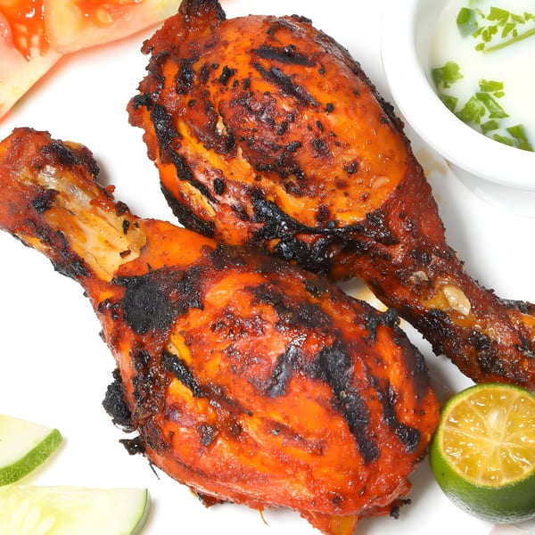Two grilled Tandoori chicken drumsticks with charred skin, served with lime, cucumber, tomato, and raita.