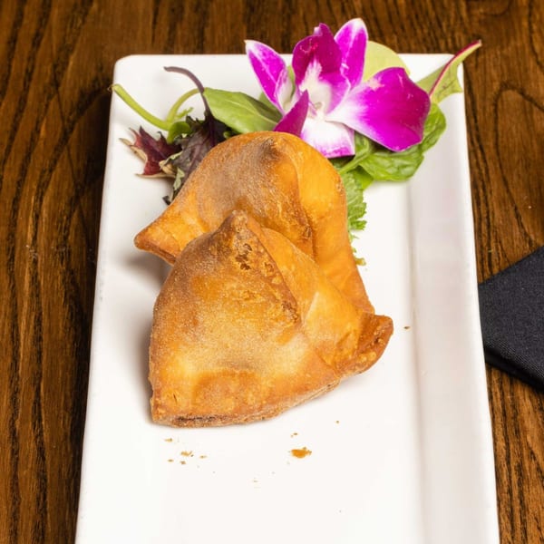 Two golden-brown samosas on a white rectangular plate, garnished with a purple orchid and greens.